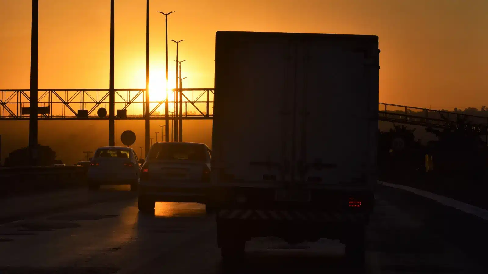 Cars and a box truck driving on a highway at sunset, silhouetted against the setting sun